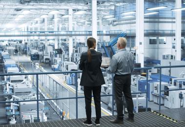 Engineers Standing with Their Back, Using Laptop Computer and Talking in a Factory Facility with Equipment Producing Modern Technical Devices for Different Industries